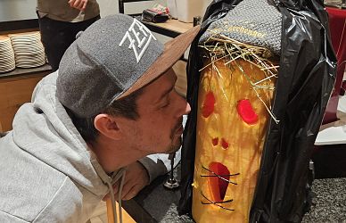Ein Mann mit grauem Basecap und Kapuzenpullover beugt sich neugierig zu einer skurrilen Figur aus Kürbis mit roten Augen und einem offenen Mund aus Stroh.