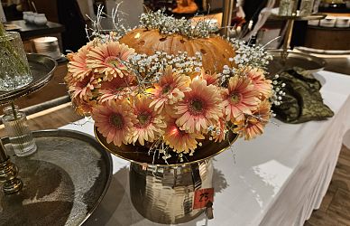 Ein Tisch in einem Geschäft, dekoriert mit Herbstblumen und einem Kürbis auf einem Metallsockel. Im Hintergrund sind weitere Dekorationsartikel zu sehen.