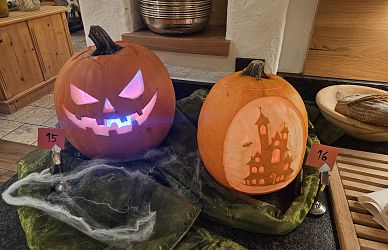 Zwei geschnitzte Halloween-Kürbisse: einer mit leuchtendem Gesicht und lila Licht, der andere mit einer Burgszene beleuchtet, auf grünen Tuch präsentiert.