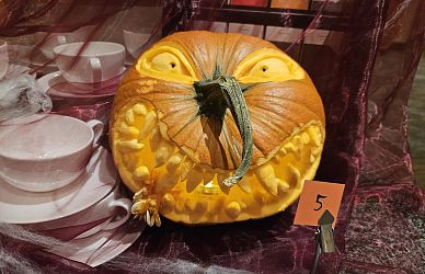 Ein geschnitzter Kürbis mit grimmigem Gesichtsausdruck steht auf einem Tisch neben weißen Schüsseln. Eine kleine 5 steht auf einem Aufsteller daneben.