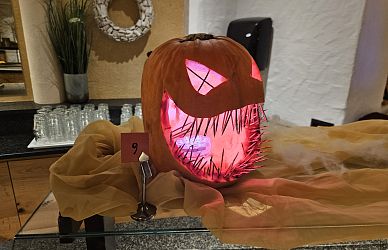 Ein geschnitzter Kürbis mit grimmigem Gesicht, leuchtend von innen, steht auf einem bedeckten Tisch. Im Hintergrund sind Gläser und Dekorationen sichtbar.