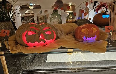 Zwei geschnitzte Kürbisse mit leuchtenden Gesichtern stehen auf einem Tisch. Im Hintergrund sind Dekorationselemente und ein elegant gedeckter Raum zu sehen.