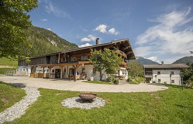 Ein traditionelles Bauernhaus in alpiner Landschaft mit Holzbalkonen, umgeben von grünen Wiesen und Bergen unter blauem Himmel mit weißen Wolken.