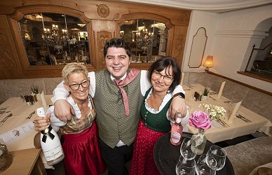 Drei Personen stehen lächelnd in einem gemütlichen Restaurant. Zwei Frauen tragen traditionelle Trachten, während der Mann in der Mitte eine Weste trägt.