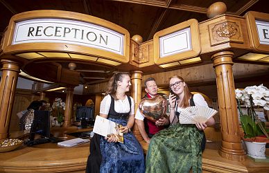 Drei Menschen in traditioneller bayerischer Tracht sitzen an einem hölzernen Empfangstresen. Sie lachen und halten verschiedene Dekorationsgegenstände.