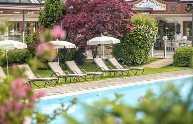 Garten mit Pool, Liegestühlen und Sonnenschirmen vor grünen Sträuchern und einem roten Baum. Im Hintergrund ein Gebäude mit großen Fenstern.