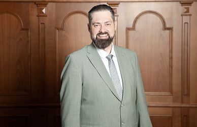 Ein Mann in einem hellgrauen Anzug steht lächelnd vor einer holzvertäfelten Wand. Er trägt ein weißes Hemd und eine gemusterte Krawatte, mit gepflegtem Bart und Haar.