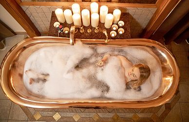 Eine Person liegt entspannt in einer modernen Kupferbadewanne, umgeben von Schaum, mit vielen brennenden Kerzen auf einem Brett am Wannenrand.