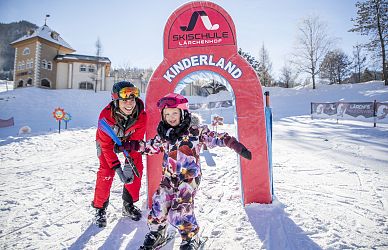 Zwei fröhliche Menschen stehen auf Skiern vor einem roten Bogen mit der Aufschrift 'Kinderland'. Im Hintergrund sind Schnee, Bäume und ein Gebäude zu sehen.