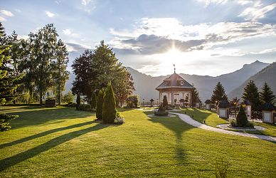 Ein gepflegter Gartenpark mit einem Pavillon in der Mitte, umgeben von Bäumen und Bergen im Hintergrund. Die Sonne scheint durch eine Wolkendecke hindurch.