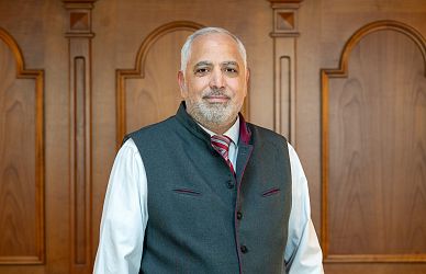 Ein Mann steht vor einer Holzwand mit dekorativen Paneelen. Er trägt eine graue Weste, ein weißes Hemd und eine rot-weiß gemusterte Krawatte.