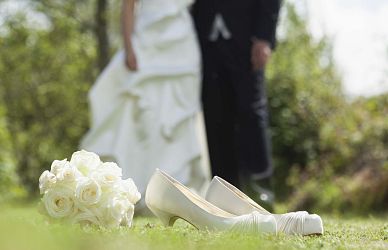 White wedding shoes and a bouquet of roses on grass; a bride and groom stand in blurred background, capturing a serene wedding moment outdoors.