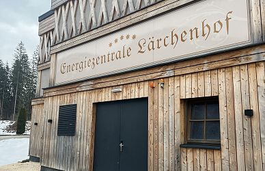Holzgebäude mit der Aufschrift „Energiezentrale Lärchenhof“, dekoriert mit Sternsymbolen. Im Hintergrund sind bewaldete Hügel und vereinzelt Schneereste sichtbar.