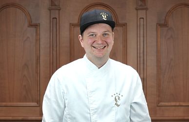 Ein Mann in weißer Kochjacke und schwarzer Kappe lächelt in die Kamera. Er steht vor einer Holzwand mit dekorativen Elementen. Sein Outfit zeigt ein Logo.