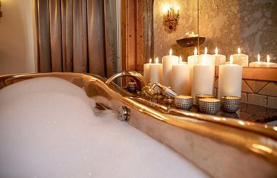 A warm, inviting bathroom scene features a foamy bathtub and several lit white candles on the side, casting a soft glow and creating a tranquil ambiance.