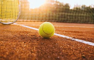 Eine Tennisball liegt auf einem sandigen Tennisplatz nahe der weißen Seitenlinie, während die Sonne im Hintergrund untergeht, was eine warme Atmosphäre schafft.