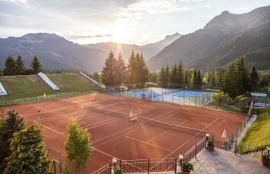 Panorama von Tennisplätzen bei Sonnenuntergang, umgeben von Bergen und Kiefern. Ein roter Sandplatz im Vordergrund und ein blauer Hartplatz im Hintergrund.