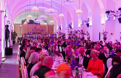 Ein festlicher Saal mit hohen, gewölbten Decken, violetter Beleuchtung und vielen Gästen in Abendgarderobe. Tische sind elegant gedeckt, Blumenarrangements schmücken den Raum.