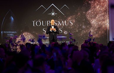 Ein Redner im Anzug steht lächelnd auf einer Bühne vor einem glitzernden Hintergrund mit der Aufschrift 