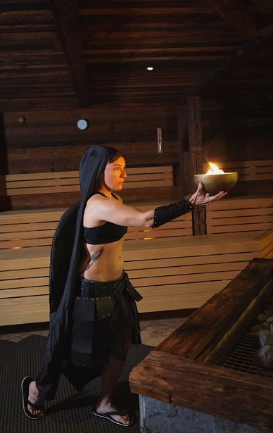 Frau in schwarzer Kleidung und Umhang hält eine brennende Schale in den Händen. Sie steht in einem Raum aus Holz, vermutlich eine Sauna oder ein rustikaler Bereich.