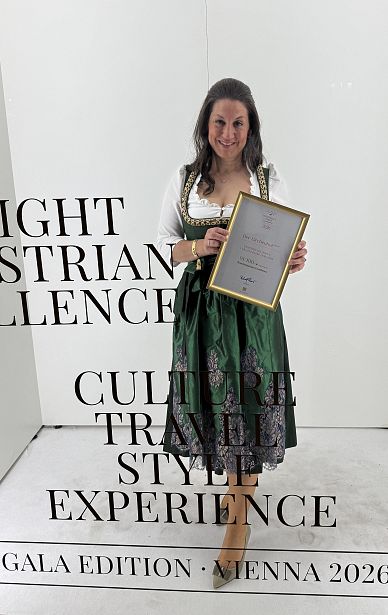 Eine Frau in traditioneller Tracht hält eine gerahmte Urkunde. Der Hintergrund ist neutral weiß, mit schwarzem Text, der teilweise lesbar ist: 