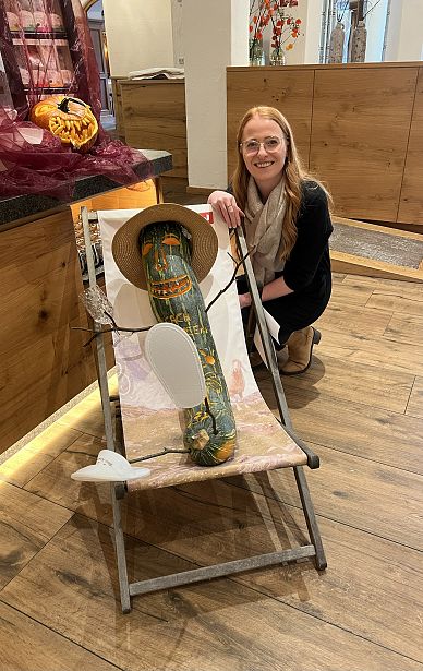 Eine Person kniet neben einem Deckstuhl, wo ein bemalter Holzpfahl mit Hut und Papierarmen sitzt, als ob es eine menschliche Figur wäre.