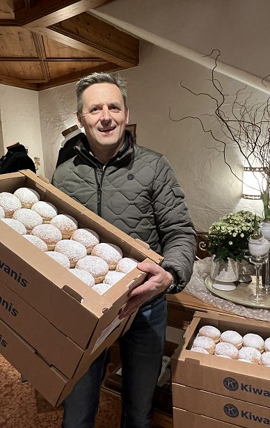 Ein Mann hält eine große Schachtel voller mit Staubzucker bestäubter Faschingskrapfen. Im Hintergrund sind Pflanzen und eine Lampe auf einem Tisch zu sehen.