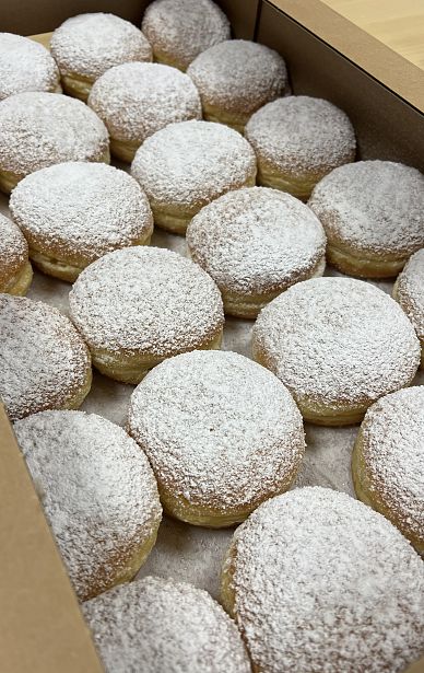 Eine Schachtel voller frischer Faschingskrapfen mit einer gleichmäßigen Schicht Puderzucker auf der Oberseite. Der Hintergrund zeigt die strukturierte, goldene Kruste der Krapfen.