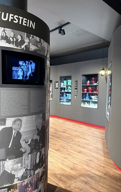 Ein Ausstellungsraum mit Holzfußboden. Links ein Monitor und Fotos an der Wand, rechts sind beleuchtete Vitrinen mit bunten Gegenständen. Eine Lampe hängt von der Decke.