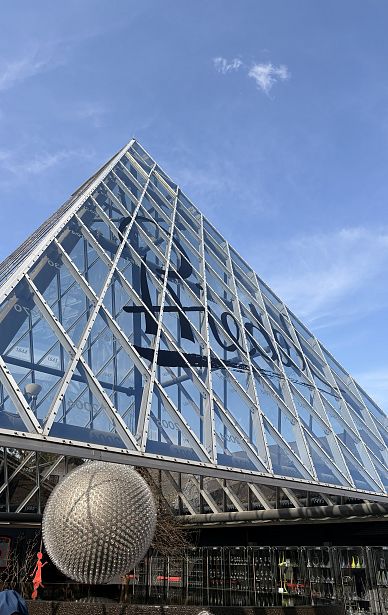 Glaspyramide mit metallischen Streben vor blauem Himmel, reflektiert Sonnenlicht. Markante Architektur mit großen, diagonal angeordneten Glasflächen. Moderne, elegante Ästhetik.