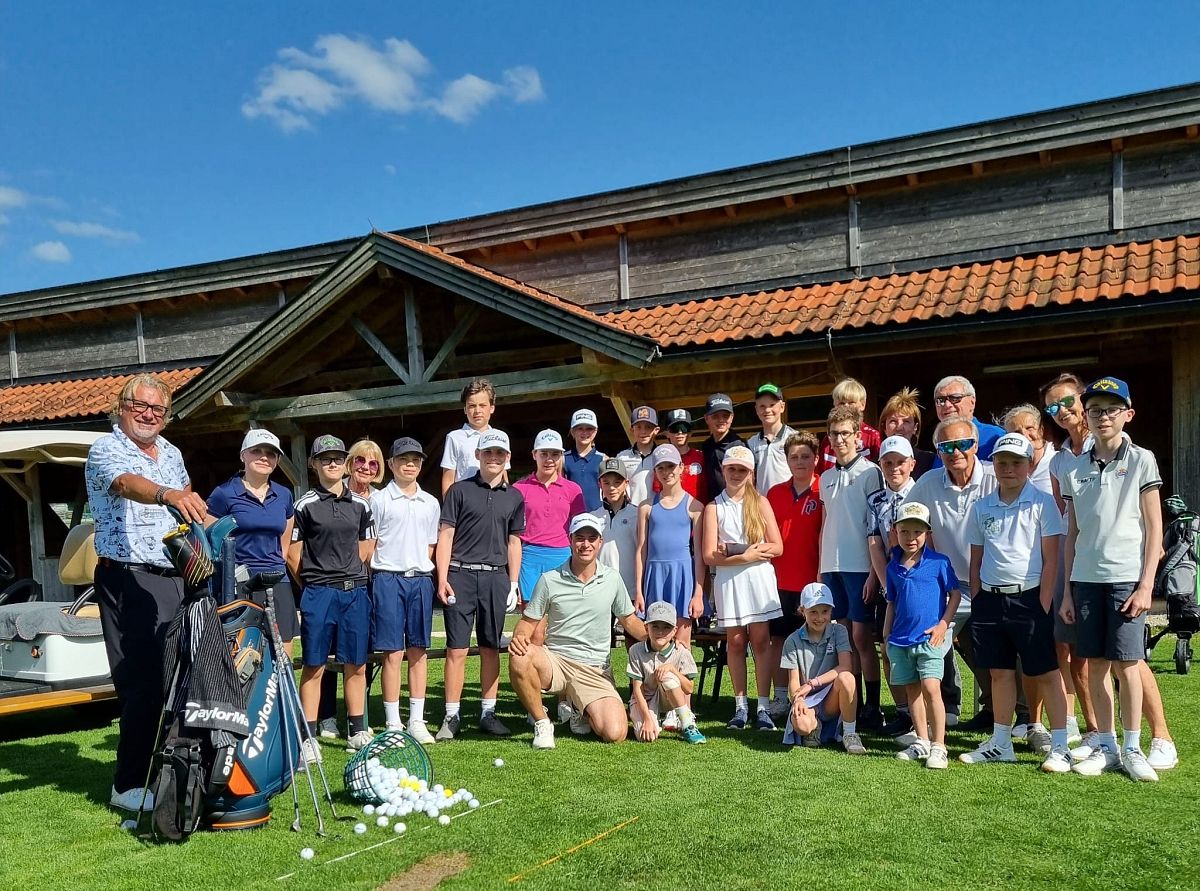 Eine Gruppe von Menschen steht vor einem Gebäude mit Holzelementen. Sie tragen Golfkleidung und lächeln in die Kamera. Golfutensilien sind ebenfalls zu sehen.