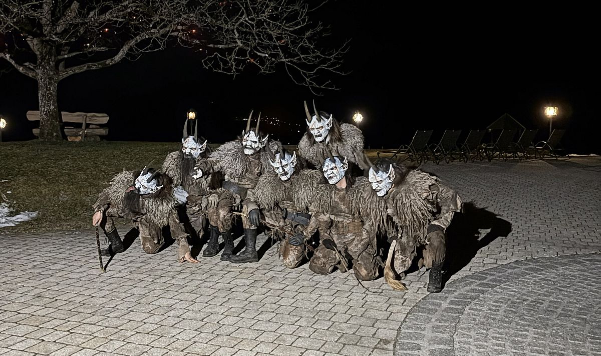 Eine Gruppe von Menschen in traditionellen Krampus-Kostümen mit Masken und Hörnern posiert nachts im Freien auf einem Pflasterweg. Im Hintergrund Bäume und Bänke.
