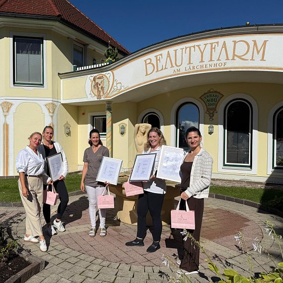Fünf Frauen stehen vor einem Gebäude mit der Aufschrift „Beautyfarm.“ Sie halten Zertifikate in den Händen und lächeln. Die Szene ist sonnig und freundlich.