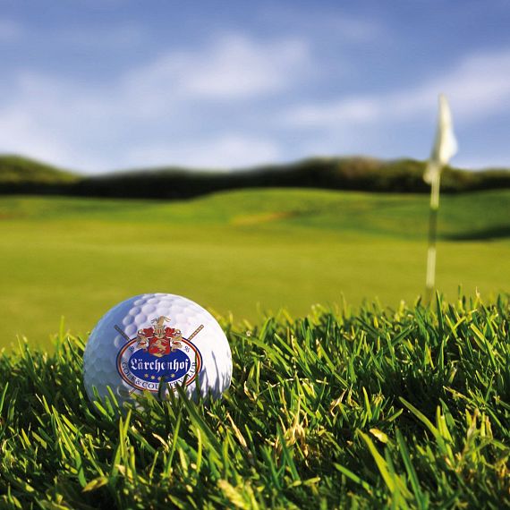 Ein Golfball liegt auf grüner Wiese, im Hintergrund ist eine Golfplatzfahne zu sehen. Der Himmel ist klar mit wenigen Wolken, was auf sonniges Wetter hinweist.