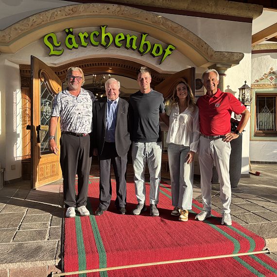 Fünf Personen stehen auf einem roten Teppich vor einem Gebäude mit dem Schriftzug „Lärchenhof“. Sie lächeln in die Kamera, während Sonnenlicht die Szene erhellt.