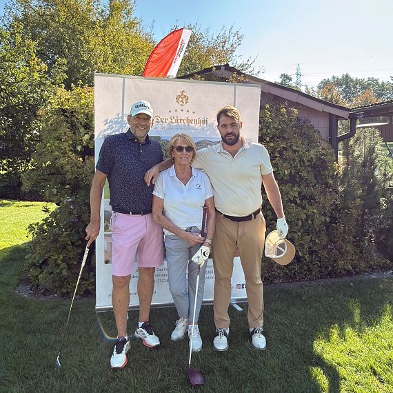 Drei Personen stehen auf einem grünen Rasen mit Golfschlägern, lächelnd vor einem Werbebanner. Die Sonne scheint hell, und sie sind leger in Golfausrüstung gekleidet.