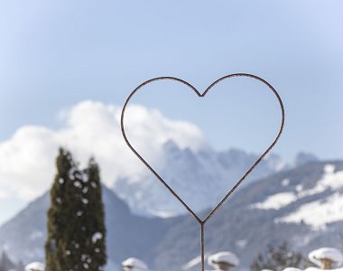 Ein herzförmiger Metallrahmen steht im Vordergrund vor einer verschneiten Berglandschaft mit klarem Himmel und Nadelbäumen, die Ruhe und Naturverbundenheit ausstrahlt.