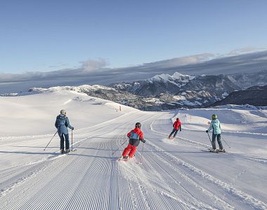 Vier Skifahrer fahren bei sonnigem Wetter auf präparierten Pisten mit malerischen schneebedeckten Bergen im Hintergrund. Perfekte Bedingungen für Wintersport.